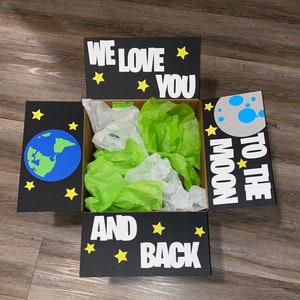 May include: A black gift box with a white background. The box has a lid with the words "We love you to the moon and back" and stars. The inside of the box is filled with green and white tissue paper.