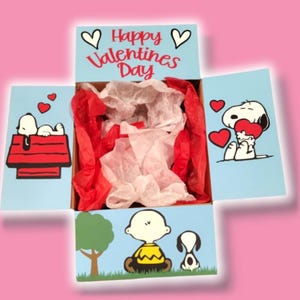 May include: A Valentine's Day gift box featuring Snoopy and Charlie Brown characters. The box is light blue with red and white tissue paper inside. The lid reads "Happy Valentines Day" with hearts.