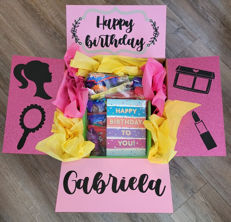 May include: Pink gift box with a glittery pink lid and a pink base. The box is open and filled with tissue paper, candy, and a card that says "Happy Birthday to You!" The box also has a black silhouette of a Barbie doll and a mirror on the left side, and a black silhouette of a lipstick and an eyeshadow palette on the right side. The bottom of the box has the name "Gabriela" written in black cursive.