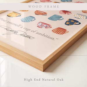 May include: A framed wall art print in a natural oak wood frame featuring a light pink background with various colorful illustrated coffee mugs and the text, "Pour yourself a cup of ambition." followed by a signature in black script lettering.