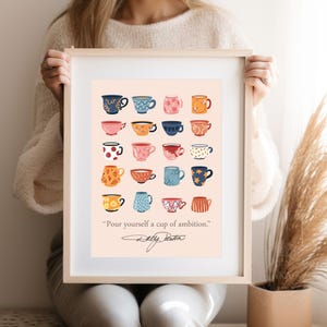 May include: A framed art print featuring a grid of twenty illustrated mugs in various colors and patterns on a light peach background. Below the mugs, the text reads, "Pour yourself a cup of ambition." followed by a signature in black ink.