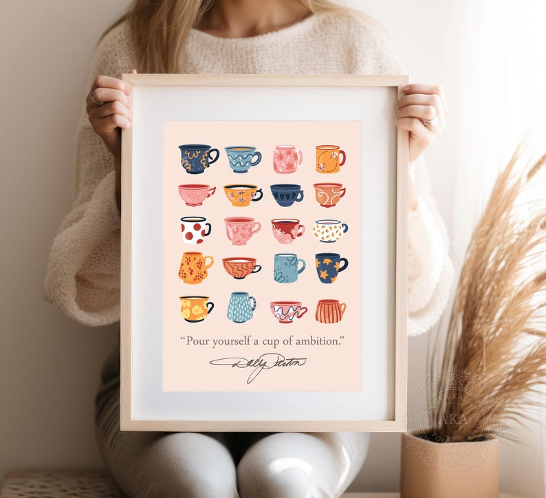 May include: A framed art print featuring a grid of twenty illustrated mugs in various colors and patterns on a light peach background. Below the mugs, the text reads, "Pour yourself a cup of ambition." followed by a signature in black ink.