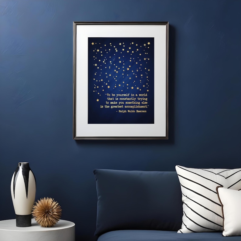 May include: A framed print with a dark blue background and gold stars. The print features a quote by Ralph Waldo Emerson: "To be yourself in a world that is constantly trying to make you something else is the greatest accomplishment."