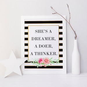 Op de afbeelding: Zwart-wit gestreepte achtergrond met gouden accenten. De tekst "She's a dreamer, a doer, a thinker." is in zwart gedrukt. Een bloemmotief met roze bloemen en groene bladeren bevindt zich onderaan de afbeelding.
