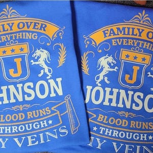 May include: Two blue t-shirts with the text "FAMILY OVER EVERYTHING" in gold and white. The shirts feature a crest with the letter "J" and the name "JOHNSON" below. The phrase "BLOOD RUNS THROUGH MY VEINS" is also displayed.