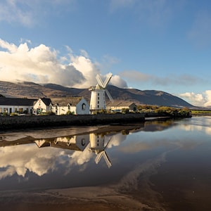 Puede incluir: Un molino de viento blanco con techo rojo se alza en la orilla de un cuerpo de agua tranquilo. El molino de viento se refleja en el agua. Hay casas y árboles en el fondo.