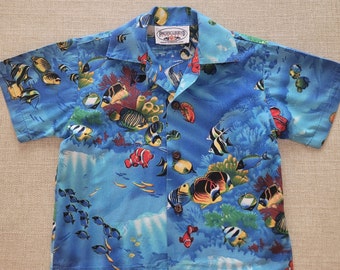 Puede incluir: Una camisa azul de manga corta con un estampado de peces tropicales coloridos. La camisa tiene un cuello abotonado y la etiqueta "Pacific Legend" es visible en la parte delantera.