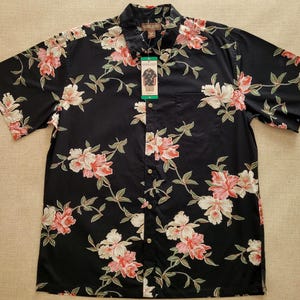 Pode incluir: Camisa preta de manga curta com um padrão floral de flores rosa e brancas e folhas verdes. A camisa tem botões, um bolso no peito e uma gola. Uma etiqueta está anexada.