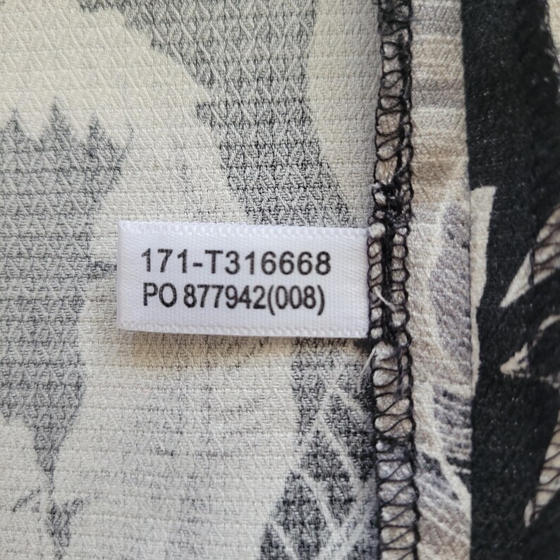 Może przedstawiać: Biała metka z czarnym napisem 171-T316668 oraz PO 877942(008) jest przyszyta do teksturowanego materiału w czarno-biały wz&oacute;r botaniczny. Krawędź tkaniny wykończona jest czarnym szwem owerlokowym z widocznymi ściegami nici.