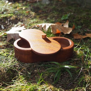 Könnte beinhalten: Eine Holzkiste mit zwei Fächern und einem Scharnierdeckel. Die Kiste ist aus dunkelbraunem Holz und hat einen kleinen Knopf auf dem Deckel. Die Kiste steht auf einem Bett aus grünem Gras und braunen Blättern.