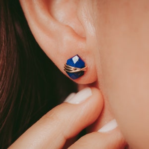 May include: A close-up of a person's ear with a blue gemstone earring. The earring is made of a gold wire wrapped around the gemstone.