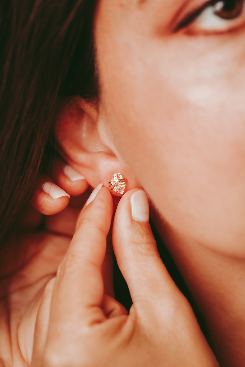 Herkimer Diamond Stud Earrings worn on the earlobe by a woman. Quartz crystals wrapped three times in gold wire for completed post earrings. The quartz crystals are transparent, colorless, and naturally faceted. They are double-terminated.