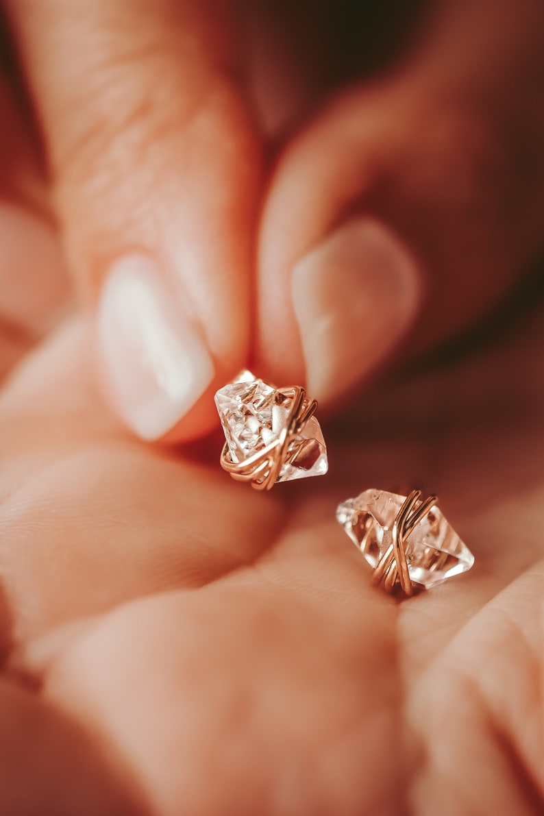 Herkimer Diamond Stud Earrings in the palm of a woman with manicured nails. Quartz crystals wrapped three times in gold wire for completed post earrings. The quartz crystals are transparent, colorless, naturally faceted, and pointed on both ends.