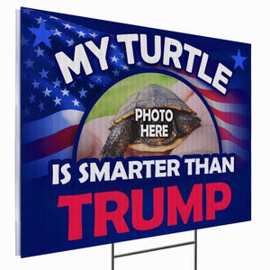 May include: A blue and white yard sign with the text "MY TURTLE IS SMARTER THAN TRUMP" in white and red. The sign features an image of a turtle with the words "PHOTO HERE" and an American flag design.