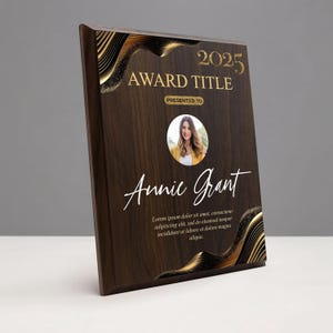 May include: A wooden award plaque with gold accents and the text "2025 AWARD TITLE" and "PRESENTED TO". It features a photo of a woman and the name "Annie Grant" in elegant script. The plaque has a dark wood grain finish.