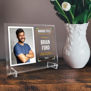 May include: A clear acrylic award with a photo of a man and text that reads "AWARD TITLE CUSTOM TEXT HERE Presented to BRIAN FORD Insert Your Custom Message or Quote Here 2024". The award is on a wooden surface.