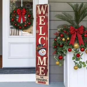 May include: A red and white wooden welcome sign with a snowman graphic and the family name. The sign is decorated with snowflakes and the word "WELCOME" in large letters. A Christmas wreath with a red bow hangs on the door.