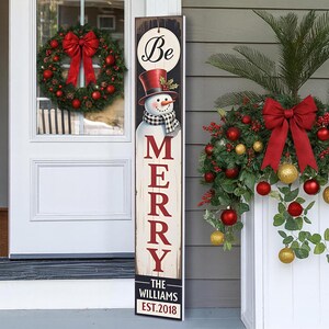May include: A wooden sign with a snowman wearing a top hat and scarf, with the word "MERRY" in red letters. The sign also includes the text "Be" and "THE WILLIAMS EST. 2018". A Christmas wreath and decorations are visible.
