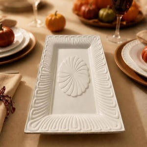 May include: A rectangular white ceramic serving platter featuring a raised, embossed fan-like pattern along the border and a central oval motif with radiating lines. The platter has a smooth, glossy finish and a clean, structured design.