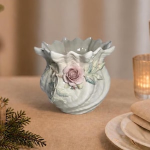 May include: A pale green ceramic vase with a scalloped, shell-like rim and a textured, swirling body. A sculpted, three-dimensional dusty pink rose with green leaves and stems is attached to the front center of the vase, adding detail to the surface.