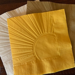 May include: Three paper napkins with a sunburst design. The napkins are in shades of white, beige, and yellow.