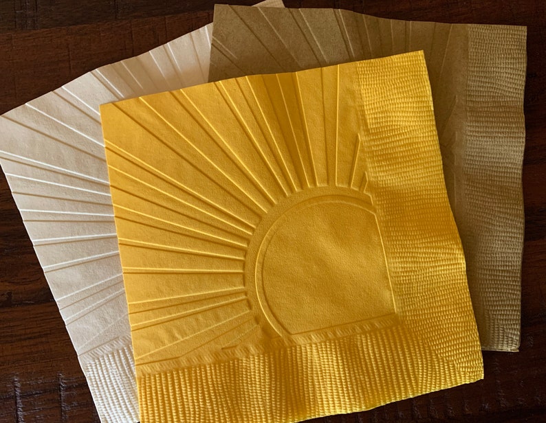 May include: Three paper napkins with a sunburst design. The napkins are in shades of white, beige, and yellow.