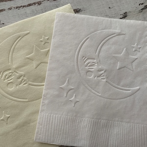 May include: Two paper napkins with a crescent moon and stars design. The napkins are white and cream colored.