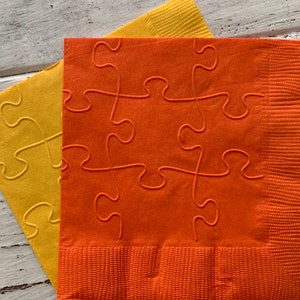 Puede incluir: Dos servilletas de papel con un diseño de piezas de rompecabezas. La servilleta de arriba es naranja y la servilleta de abajo es amarilla.
