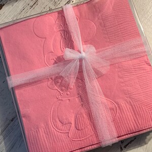 May include: Pink paper napkins featuring an embossed Minnie Mouse design, neatly stacked in a clear plastic box. The napkins are tied with a white tulle ribbon and bow, suitable for gifting.
