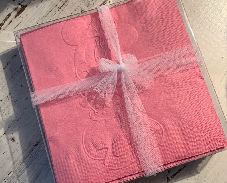 May include: Pink paper napkins featuring an embossed Minnie Mouse design, neatly stacked in a clear plastic box. The napkins are tied with a white tulle ribbon and bow, suitable for gifting.