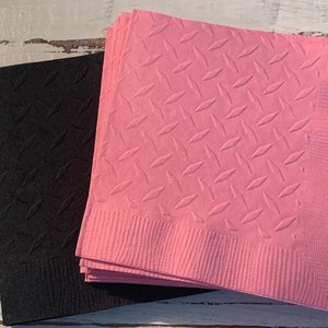 May include: A stack of pink paper napkins with a diamond pattern embossed on the surface. Next to the pink napkins is a stack of black paper napkins with the same diamond pattern embossed on the surface.
