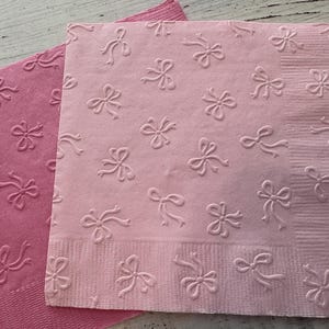 May include: Two pink paper napkins with embossed bow patterns. One napkin is a darker shade of pink, while the other is a lighter shade. The napkins are square and have a textured surface.