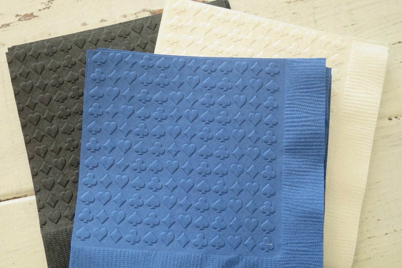 Puede incluir: Tres servilletas de papel con un patr&oacute;n en relieve de palos de cartas de juego. Las servilletas son de color negro, azul y blanco.