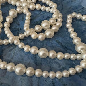 Puede incluir: Varias hebras de collares de perlas falsas de diferentes tamaños y longitudes. Las perlas son de color blanco crema y tienen un acabado brillante. Los collares están dispuestos sobre una superficie de tela azul.