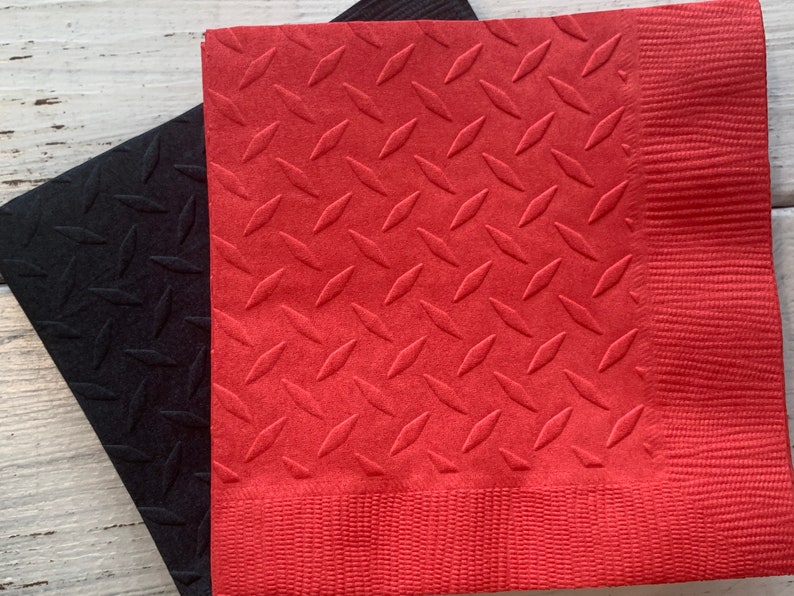 May include: Two red and black paper napkins with a diamond pattern embossed on the surface. The napkins are folded and stacked on top of each other.