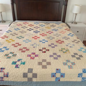 May include: A cream-colored quilt with a repeating pattern of colorful patchwork blocks. The blocks are in shades of red, blue, purple, and green. The quilt is on a dark brown bed frame with white nightstands and lamps.