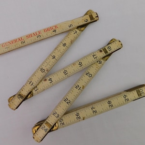 May include: An antique wooden folding ruler, measuring up to 30 inches. The ruler is cream-colored with black markings and brass joints. The text "GENERAL SHALE BRICK" is printed in red on one section. A vintage measuring tool.