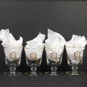 May include: A set of four clear glass goblets with short stems and circular bases. Each glass features a gold-colored decorative emblem on the front, consisting of the letters RC surrounded by an ornate frame. White tissue paper fills each glass.