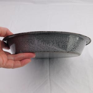 May include: A gray enamelware pie plate with a speckled pattern. The plate has a slightly raised rim and two small handles. The color is a muted gray with darker gray speckles, giving it a vintage look.