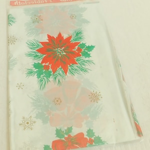 May include: A rectangular Christmas tablecloth packaged in a clear plastic sleeve. The white fabric features a repeating pattern of red poinsettia flowers, green holly leaves, pine branches, gold snowflakes, and red bows with bells.