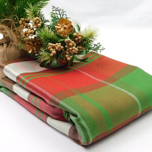 May include: A folded fabric textile featuring a plaid pattern in shades of red, green, and off-white. The fabric is accented by a decorative arrangement of green pine needles, gold-toned berries, and a small pinecone wrapped in burlap.