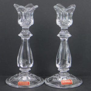 May include: Two clear crystal candlesticks with a tulip-shaped top and a flared base. The candlesticks have a detailed, elegant design and are labeled with the brand "GORHAM" on the base. The background is a solid black.