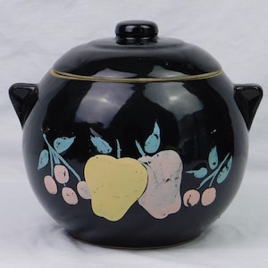 May include: Black ceramic cookie jar with a lid and two handles. The jar is decorated with a hand-painted design of apples and berries in pink, yellow, and blue.