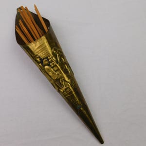 May include: A vintage brass toothpick holder with a conical shape. The holder is adorned with embossed figures and filled with wooden toothpicks. The brass has a warm, aged patina, and the design suggests a decorative, functional item.