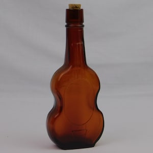 May include: A brown glass bottle shaped like a violin, with a cork stopper. The bottle has a rounded body and a long neck. The glass has a slight amber tint.