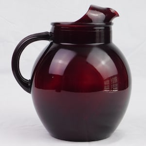 May include: A deep red glass pitcher with a rounded body, a curved handle, and a flared spout. The pitcher has a glossy finish, reflecting light. The handle is thick and curved, and the spout is slightly wavy. The pitcher is likely used for serving beverages.