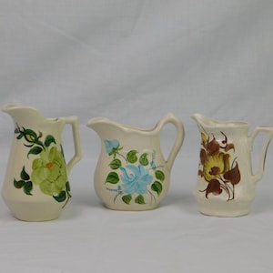 May include: Three cream-colored ceramic pitchers with floral designs. The left pitcher features a large green flower, the center pitcher displays a light blue flower, and the right pitcher shows a brown and yellow floral pattern with dark leaves.
