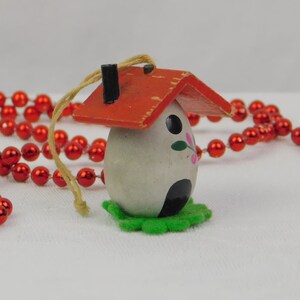 May include: A small egg-shaped ornament featuring a red painted wooden roof with a black chimney, a white body with black and pink floral accents, a black arched doorway, and a green felt base, suspended by a tan string with red bead garland.