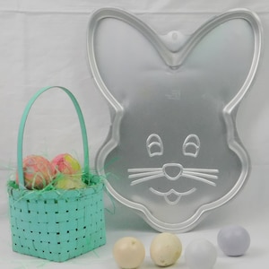 May include: A silver-colored bunny-shaped cake pan with a basket of decorated eggs and several decorative eggs. The basket is light blue and filled with green decorative grass. The eggs are pastel colors.