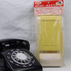 May include: A black rotary telephone with a circular dial and a yellow Akay Memo-Master note dispenser in its original plastic packaging, featuring a red header card with the text MEMO-MASTER and 3.150 square inches of writing surface.
